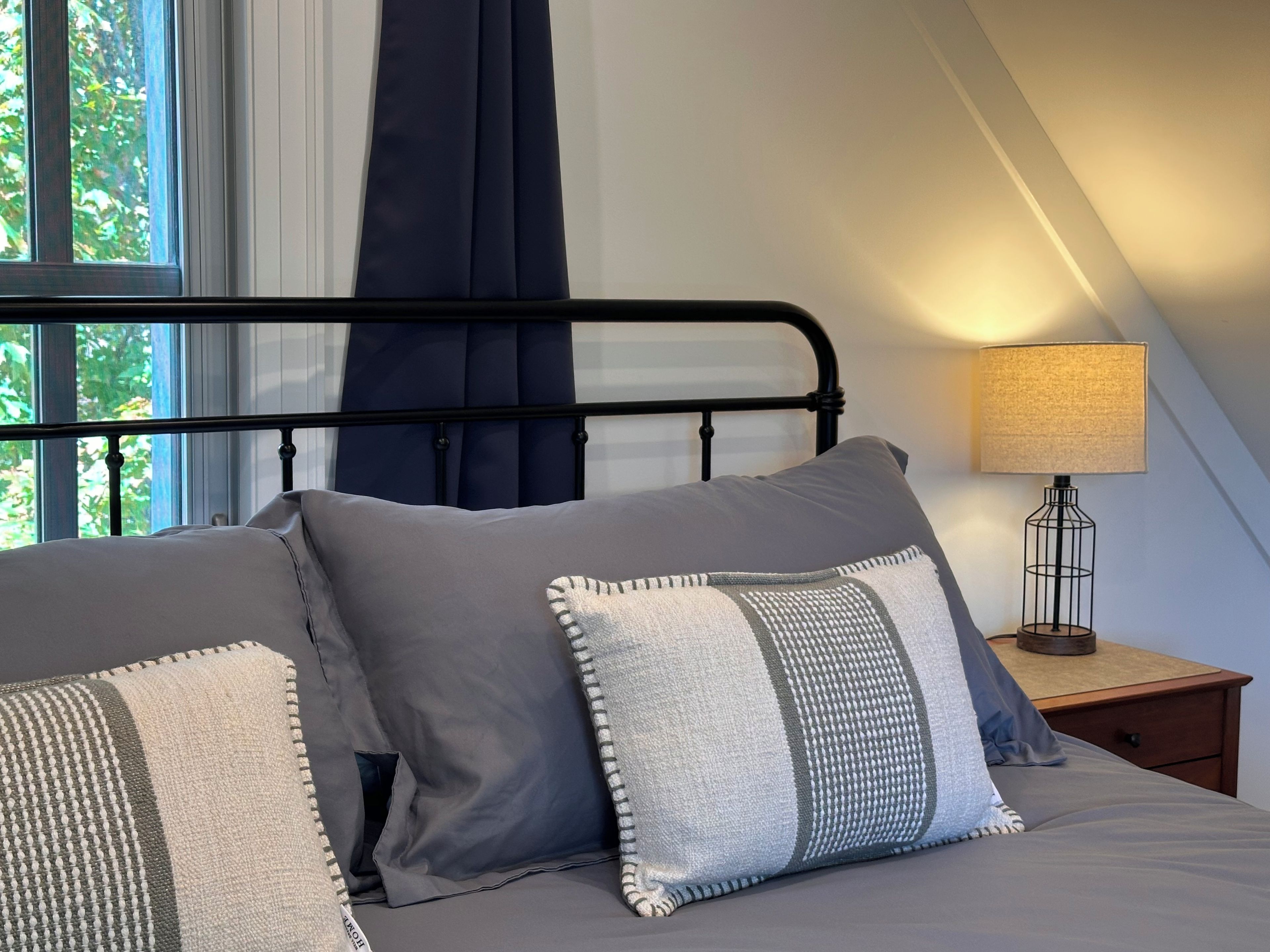 Ein Schlafzimmer mit Bett, Kissen, einem Nachttisch, einer Lampe und einem Fenster mit Vorhängen.