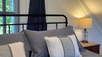 Ein Schlafzimmer mit Bett, Kissen, einem Nachttisch, einer Lampe und einem Fenster mit Vorhängen.