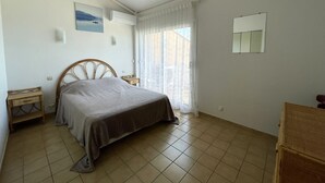 A bedroom with a bed, a bedside table, a nightstand, a window with curtains, and a wall-mounted air conditioning unit. - T3 Apartment in Banyuls-sur-Mer, terrace and private parking (Banyuls-sur-Mer)