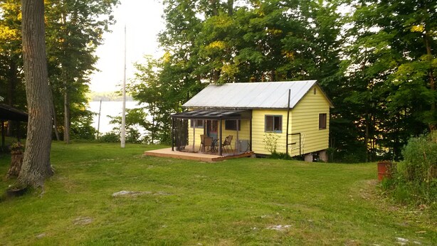 Une petite cabane jaune avec un porche et un patio couvert, entourée d’arbres et d’une pelouse.