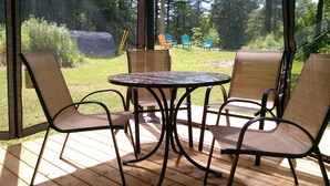 Une terrasse couverte avec une table ronde et quatre chaises, entourée d’une terrasse en bois et donnant sur un espace verdoyant agrémenté d’arbres et de mobilier d’extérieur.