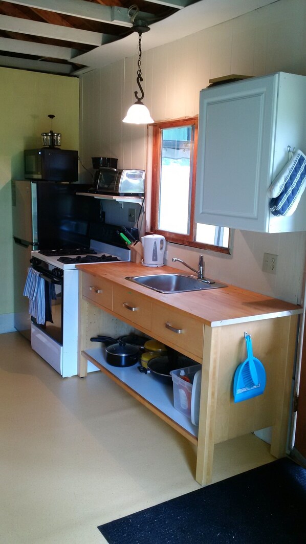 Cocina pequeña con estufa, microondas y fregadero. Una ventana proporciona luz natural y un colgador de ollas sobre el fregadero.