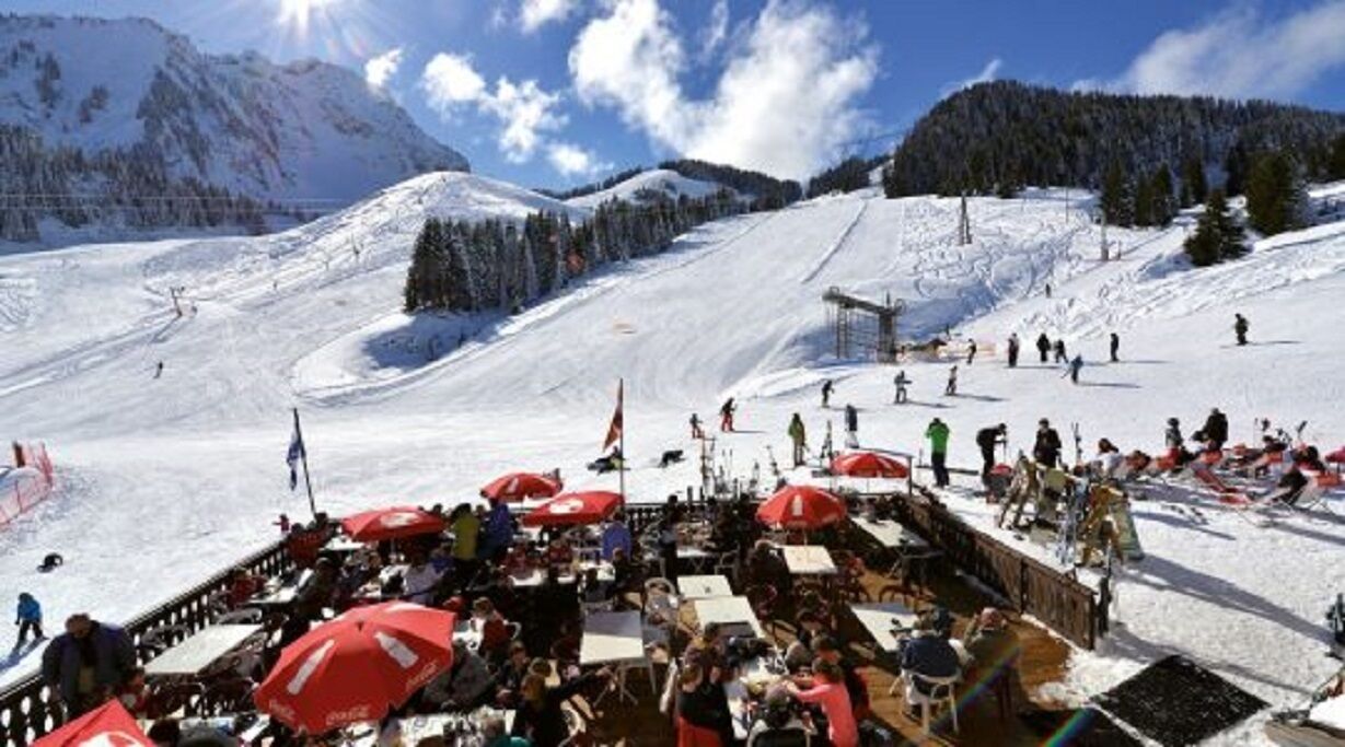 Une station de ski dotée d’un télésiège, de pistes enneigées et d’un espace de restauration avec des parasols rouges.