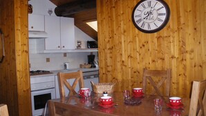 Un intérieur en bois avec une table à manger dressée pour quatre personnes, une cuisine en arrière-plan et une horloge murale.