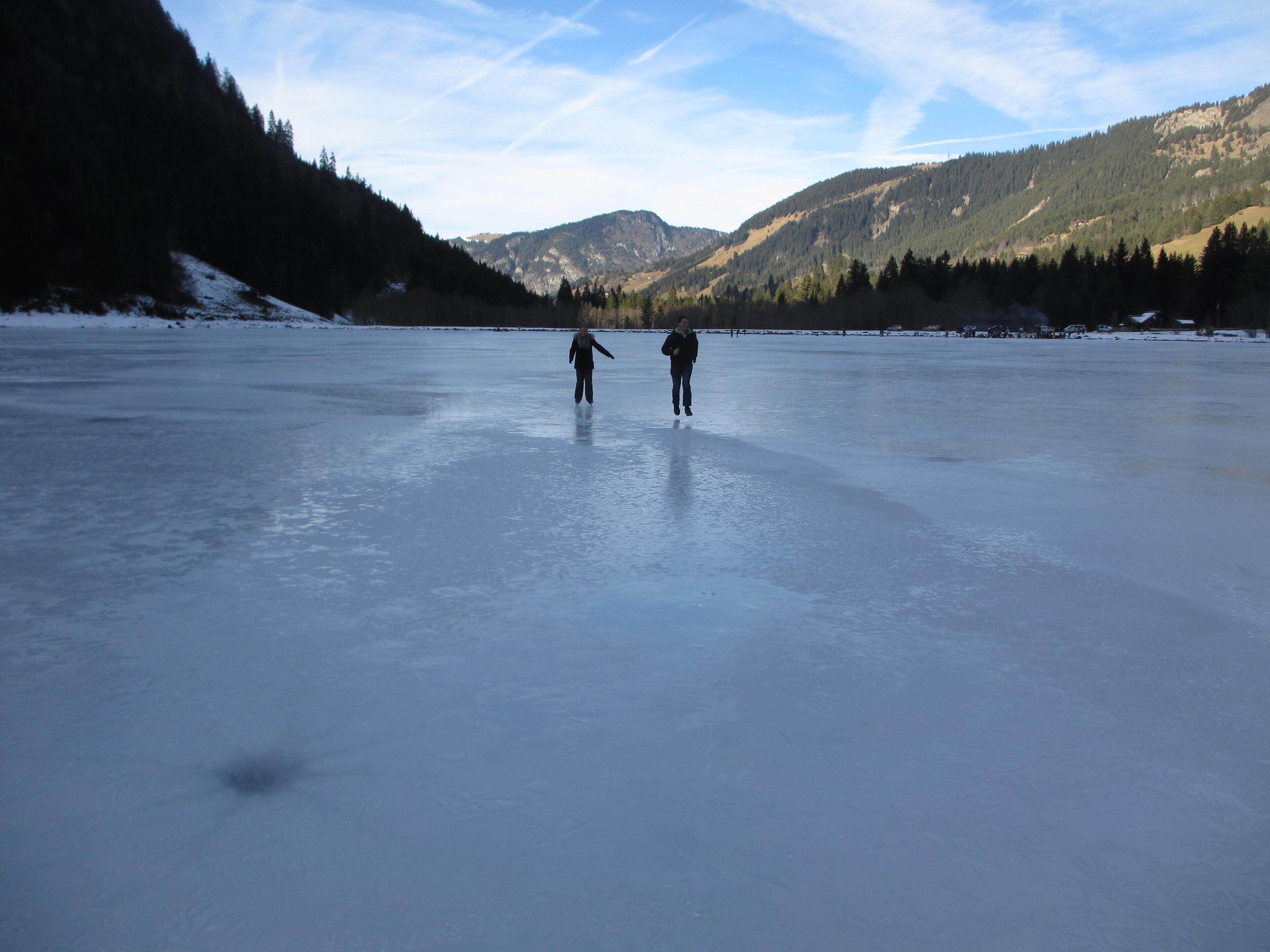 A frozen lake with two people walking on it, surrounded by forested hills.