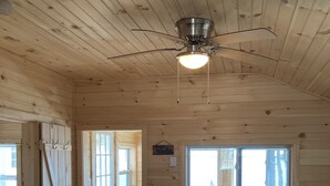 A wooden interior with a ceiling fan, a television on a stand, a sofa, and a window with a view. - TWO Waterfront Camps on Sebago Lake (Standish)