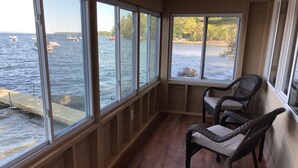 A room with large windows overlooking a body of water, wooden flooring, and wicker chairs. - TWO Waterfront Camps on Sebago Lake (Standish)