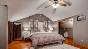 Une chambre avec un plafond en pente, un lit surmonté d’un tapis blanc, deux tables de chevet avec des lampes et un ventilateur de plafond.