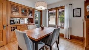 Un coin repas avec une table et des chaises en bois, un luminaire suspendu et une vue sur l’extérieur par une fenêtre agrémentée de rideaux.