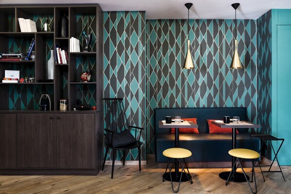 Ein modernes Interieur mit einem dunklen Holzschrank, tealfarbenem Wandpapier mit Muster, einem blauen Sofa mit roten Kissen und zwei schwarzen Stühlen mit gelben Hocker.