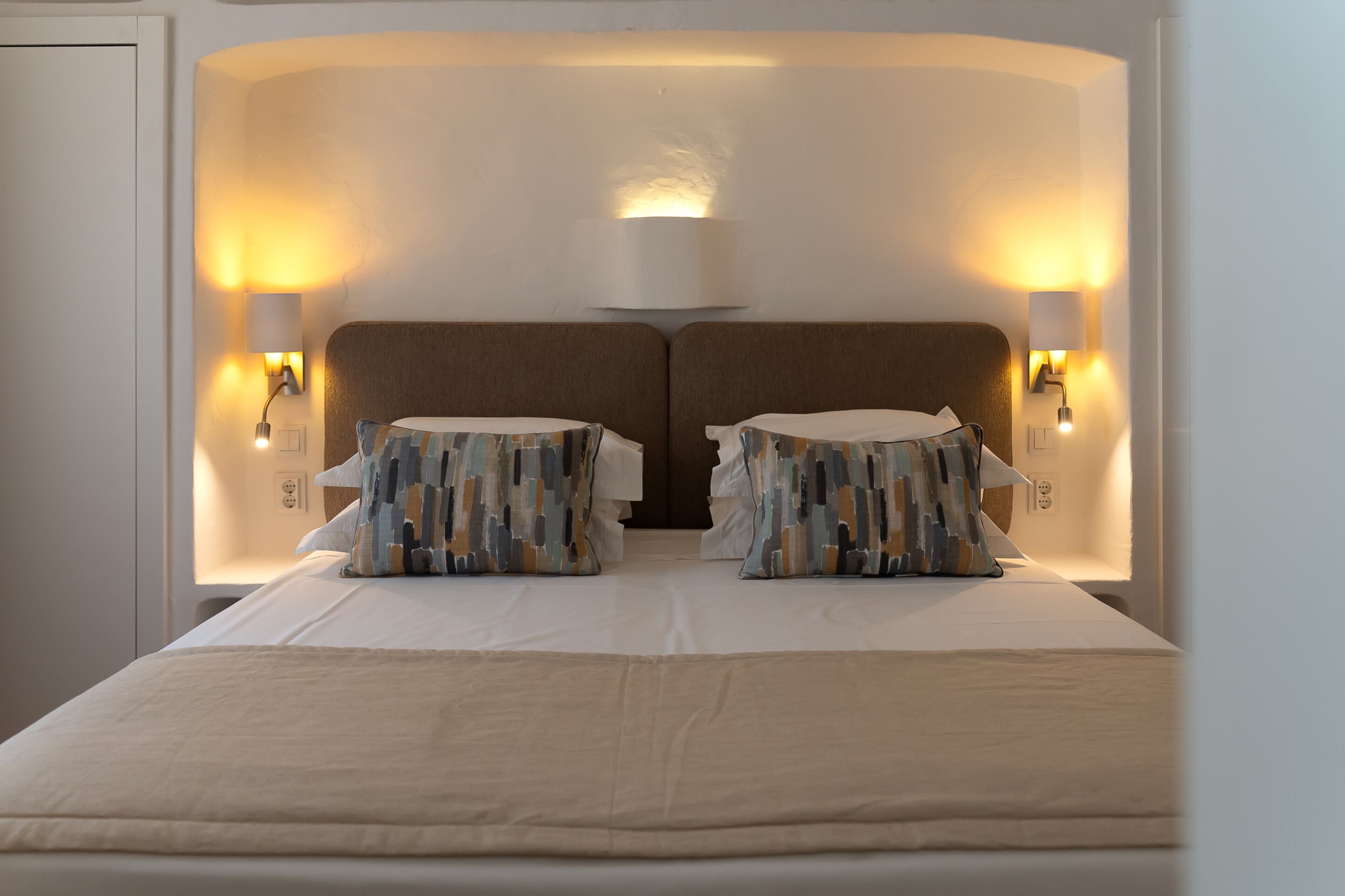 A double bed with a beige bedspread, two patterned pillows, and two wall-mounted lamps on either side.
