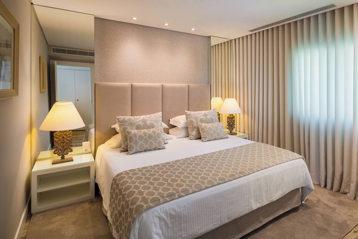 A hotel room with a large bed, two bedside lamps, a headboard, and a window with curtains.