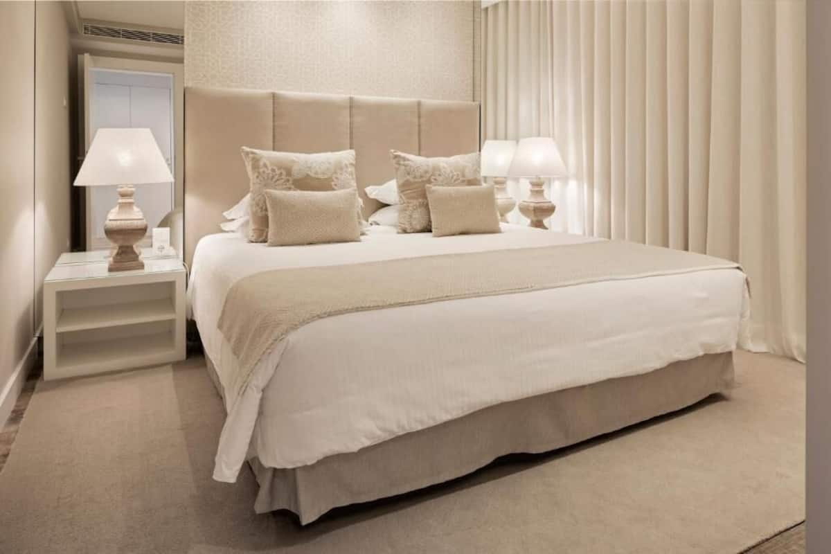 A large bed with a headboard, two bedside tables with lamps, a mirror, and a window with blinds.