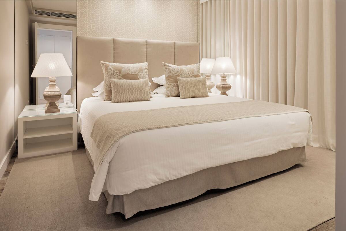 A large bed with a headboard, two bedside tables with lamps, and a window with vertical blinds.