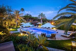 Ein Swimmingpool, umgeben von Liegestühlen und Sonnenschirmen, mit Palmen und gepflegter Bepflanzung.