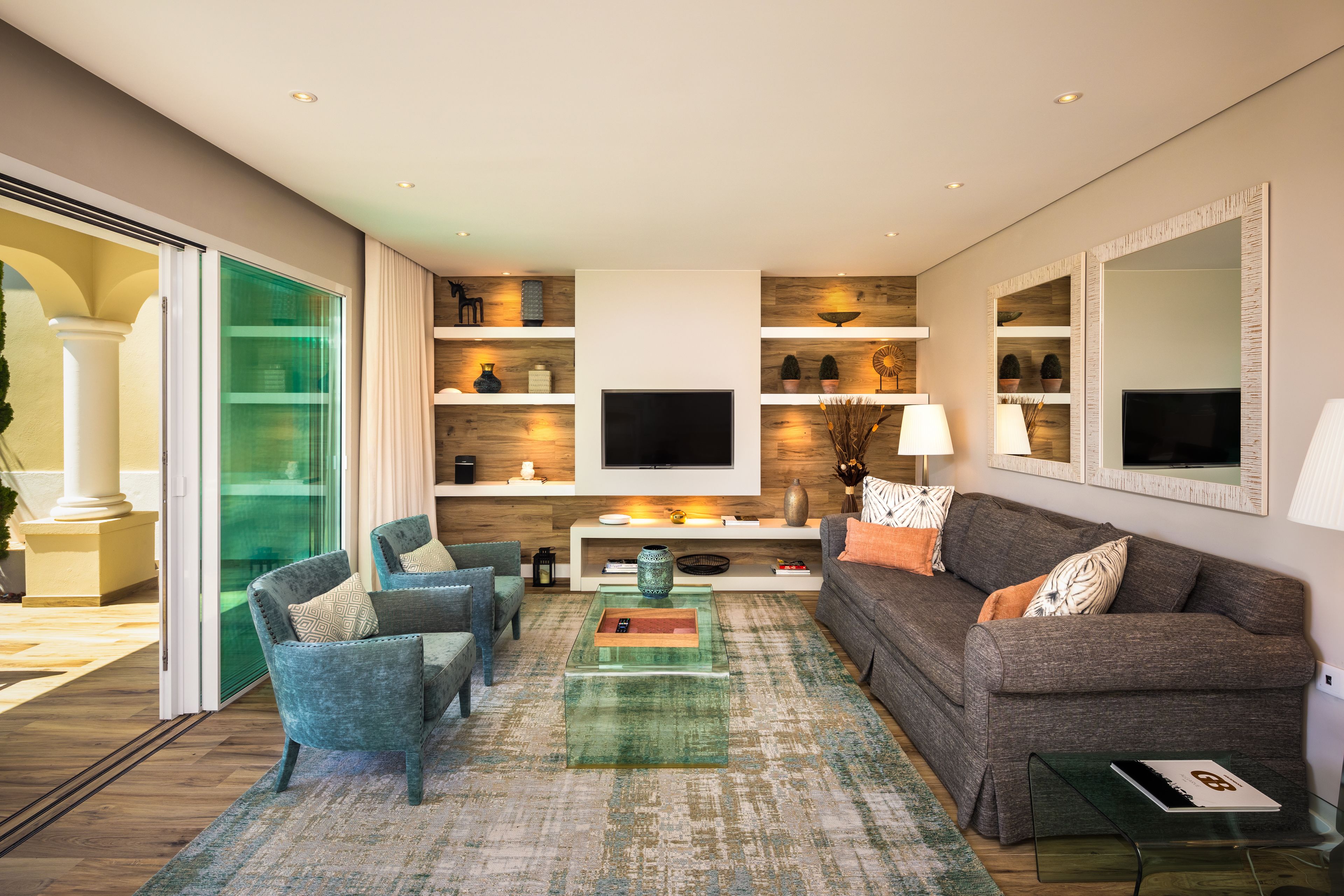 A modern living room with a sofa, chairs, a coffee table, and a TV. There are shelves with decorative items and a large mirror on the wall.