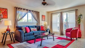 Un salon avec un canapé bleu, un fauteuil rouge et une table basse. Il y a deux fenêtres avec des rideaux, un ventilateur de plafond et un tapis rouge.