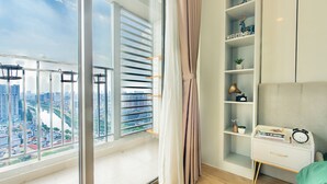 A modern bedroom with a city view, a white bedside table, a shelf with decorative items, and a window with blinds. - The Grand Saigon Apartment - City Centre (Ho Chi Minh City)