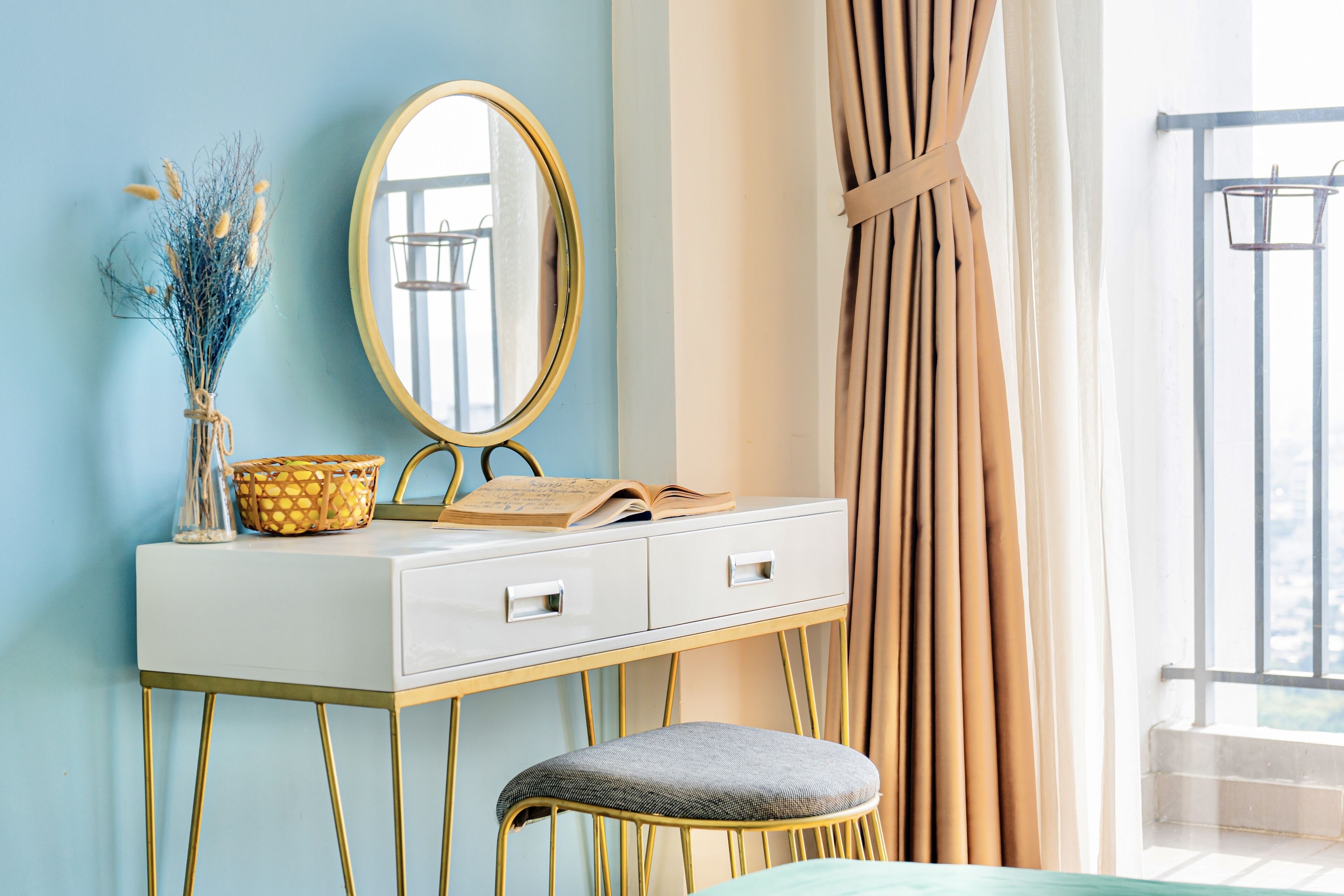 A white vanity with gold legs, a mirror, and a book. There is a stool with a cushion and a vase with dried flowers.