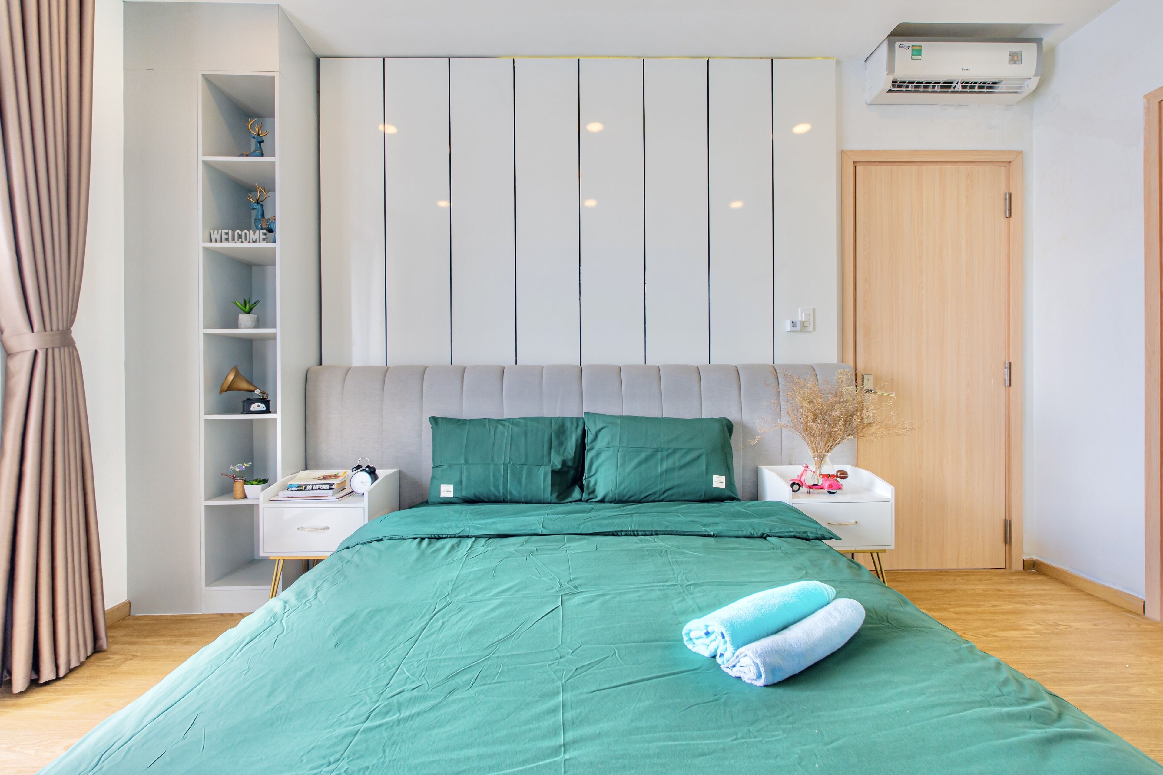 A neatly arranged bedroom with a green bedspread, a white bedside table with a lamp, a shelf with decorative items, and a wooden door.