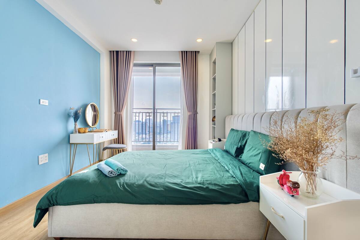 Ein Schlafzimmer mit Bett, Nachttisch, Kommode und Blick auf die Stadt durch große Fenster.