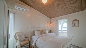 Une chambre avec un plafond en bois, un lit avec une literie blanche, une chaise avec un coussin, un luminaire suspendu et une fenêtre donnant sur un beau paysage.