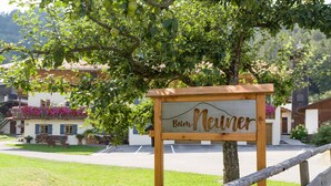 A wooden sign with the text "Bäum Neuner" in front of a building with a balcony, surrounded by greenery and a wooden fence. - Apartment Wildbarren, 55sqm, balcony (Oberaudorf)