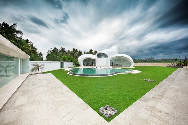 A modern pool area with a unique architectural design, a well-maintained lawn, and a clear sky. - The Cloud Unique Beachfront Villa (Tabanan)
