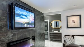 Ein modernes Wohnzimmer mit einem Flachbildfernseher an einer dunkel gefliesten Wand, einem Kamin mit sichtbarem Feuer, einem dunklen Sofa mit gemusterten Kissen und einem gerahmten Kunstwerk an der Wand.