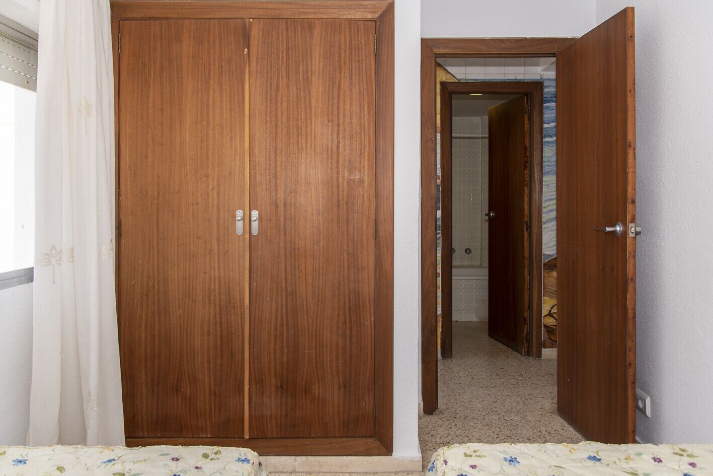 A bedroom with a wooden wardrobe, a bed with floral bedding, and a bathroom visible through an open door.