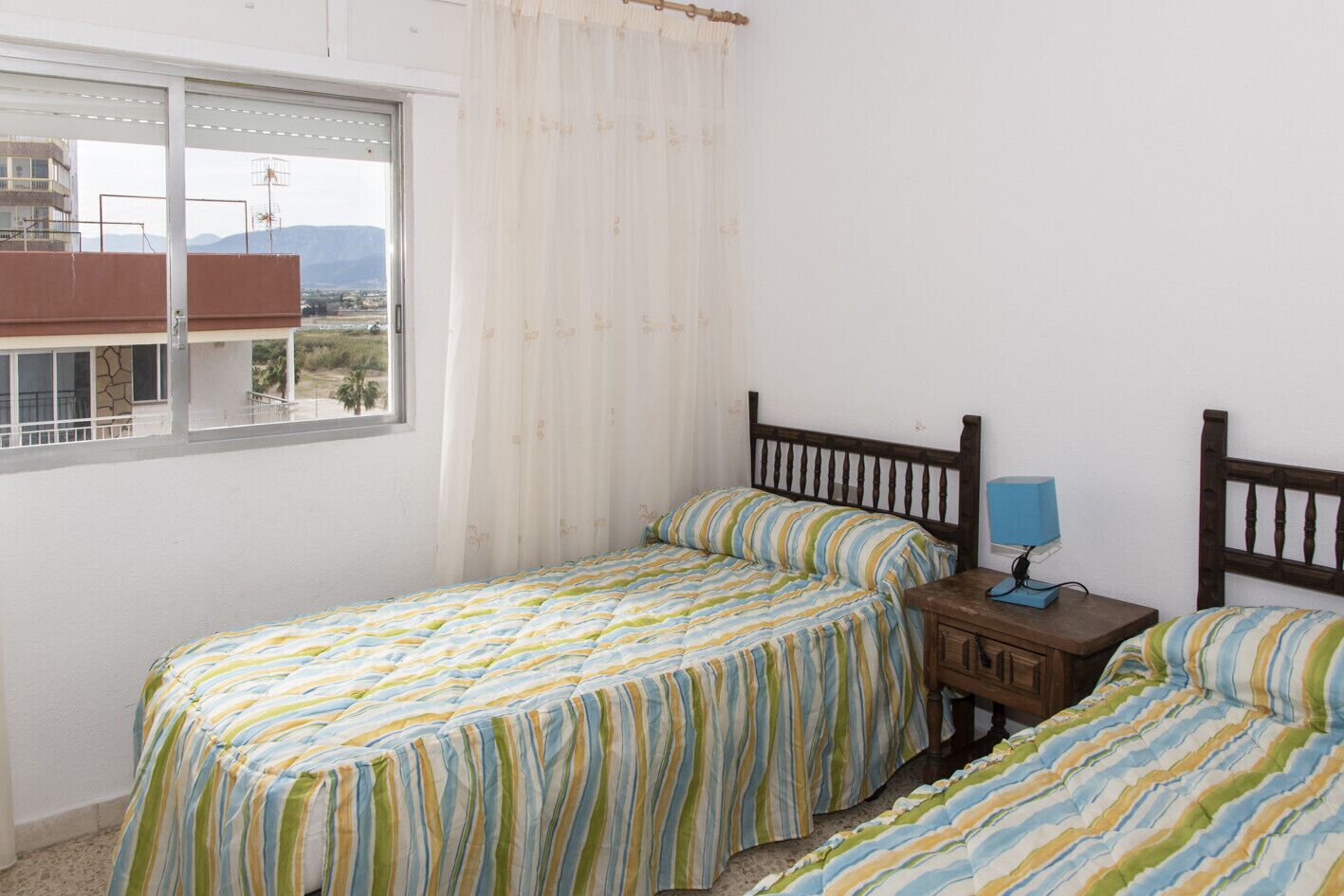 A bedroom with two single beds, a nightstand with a lamp, and a window overlooking a balcony.