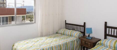 A bedroom with two single beds, a nightstand with a lamp, and a window overlooking a balcony.