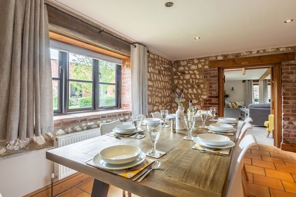 A dining area with a long wooden table set for six, a stone wall, and a view of greenery outside.