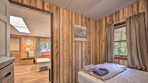 A room with wooden panel walls, a bed with a white mattress and a folded towel, a wooden dresser, a desk with a lamp, a clock, and a window with curtains. - ‘John Wayne' Apartment - Deck, BBQ, Horses On-Site (Huntingdon)