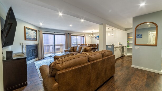 A spacious living room with a brown leather sofa, a flat-screen TV, and a fireplace. There is a dining area with a table and chairs, and a kitchen area with a countertop and cabinets.
