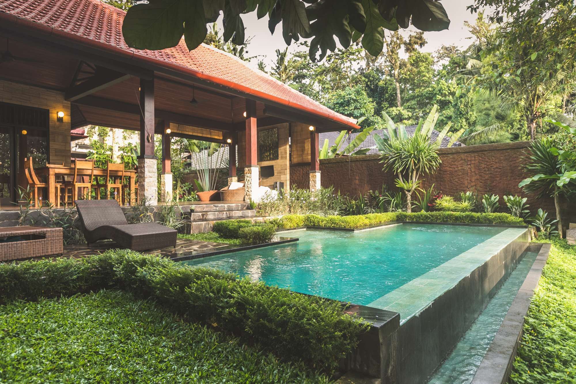 A pool area with a covered seating area, lush greenery, and a stone wall.