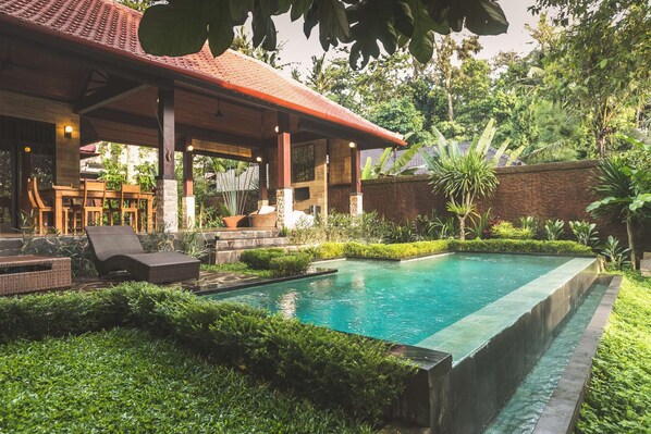 Ein Poolbereich mit überdachter Lounge-Ecke, üppiger Bepflanzung und einer Steinsäule.