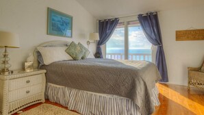 Un dormitorio con cama, mesitas de noche, lámpara, un cuadro enmarco y vista al mar a través de una ventana con cortinas.