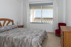 A bedroom with a bed, a nightstand, a red chair, a wooden dresser, and a window with a view of the sea.