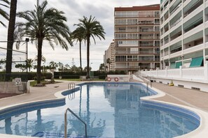 A swimming pool with a clear blue water, surrounded by a white fence, palm trees, and a multi-story building with balconies.