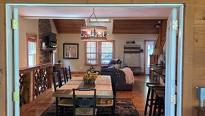 A spacious dining area with a high ceiling and a chandelier, wooden walls, and a view into a living room with a sofa and a television. - Peaceful Mountain Cabin (Sapphire)