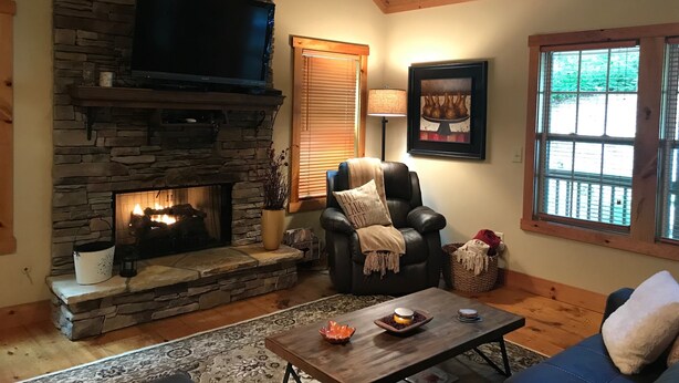 A cozy living room with a stone fireplace, a leather sofa, a coffee table, and a television mounted on the wall. - Peaceful Mountain Cabin (Sapphire)