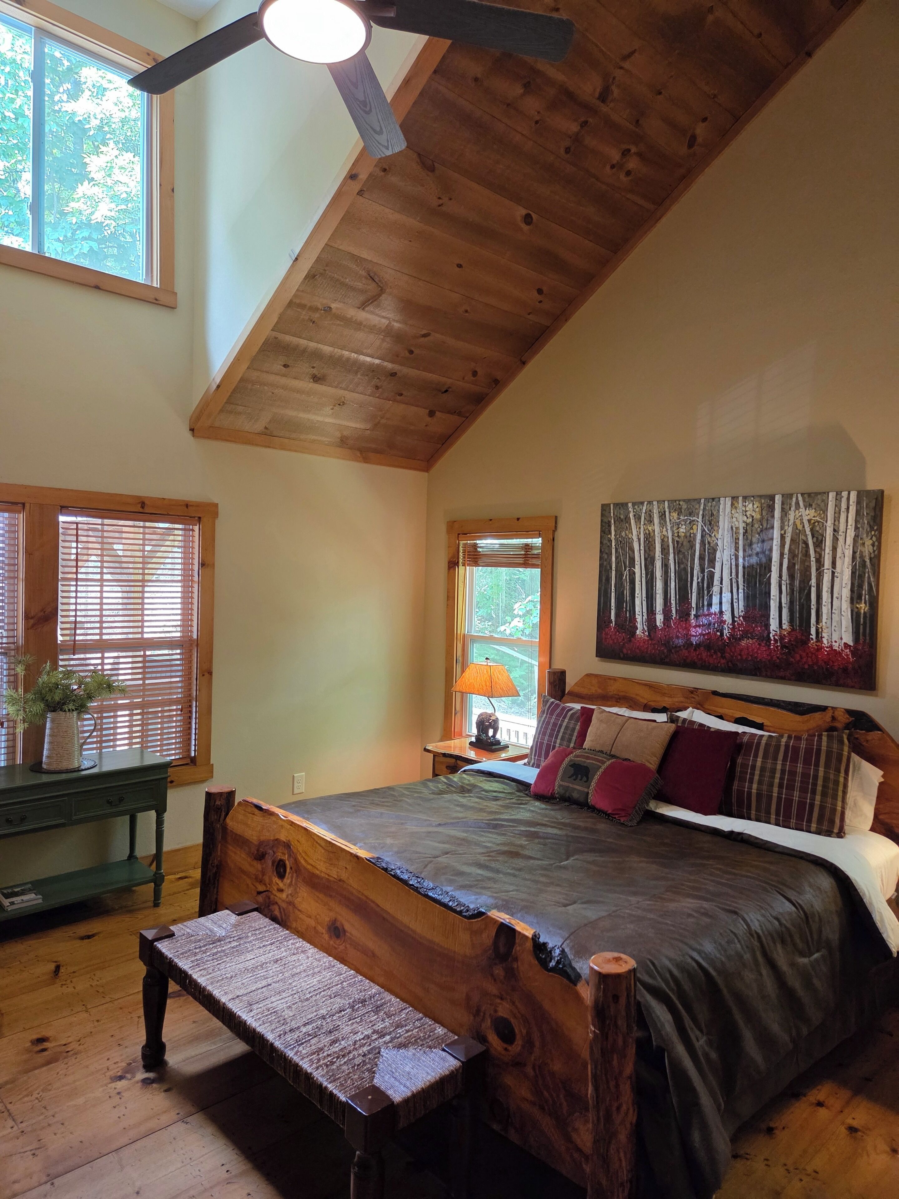 A bedroom with a sloped ceiling, wooden bed frame, a bench, a bedside table with a lamp, and a painting of trees.
