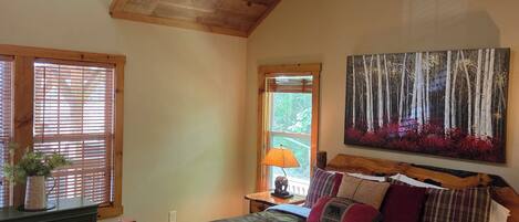 A bedroom with a sloped ceiling, wooden bed frame, a bench, a bedside table with a lamp, and a painting of trees.