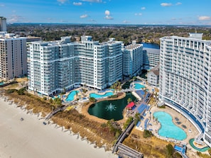A high-rise hotel with multiple balconies, a central swimming pool, and a beach area.