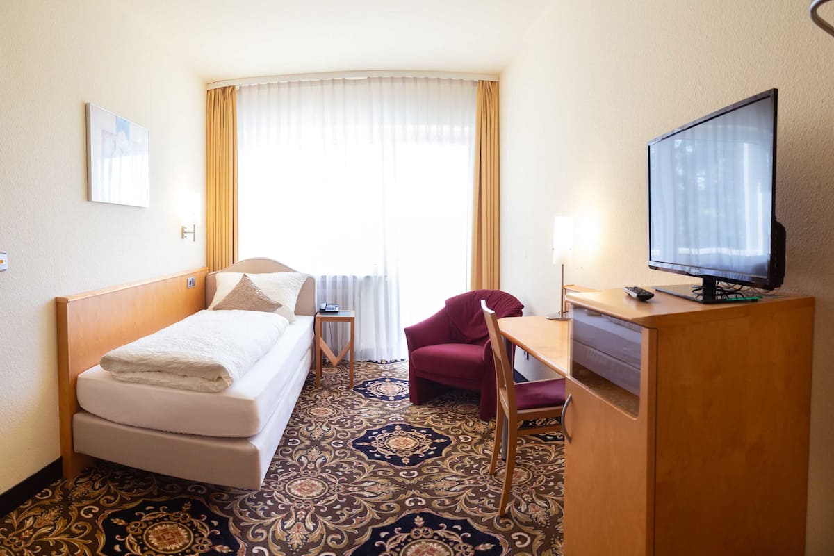 Quarto de hotel com cama, escrivaninha com televisão, cadeira, mesa pequena e tapete estampado.