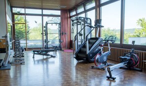 Academia com diversos equipamentos de ginástica, janelas amplas e piso de madeira.