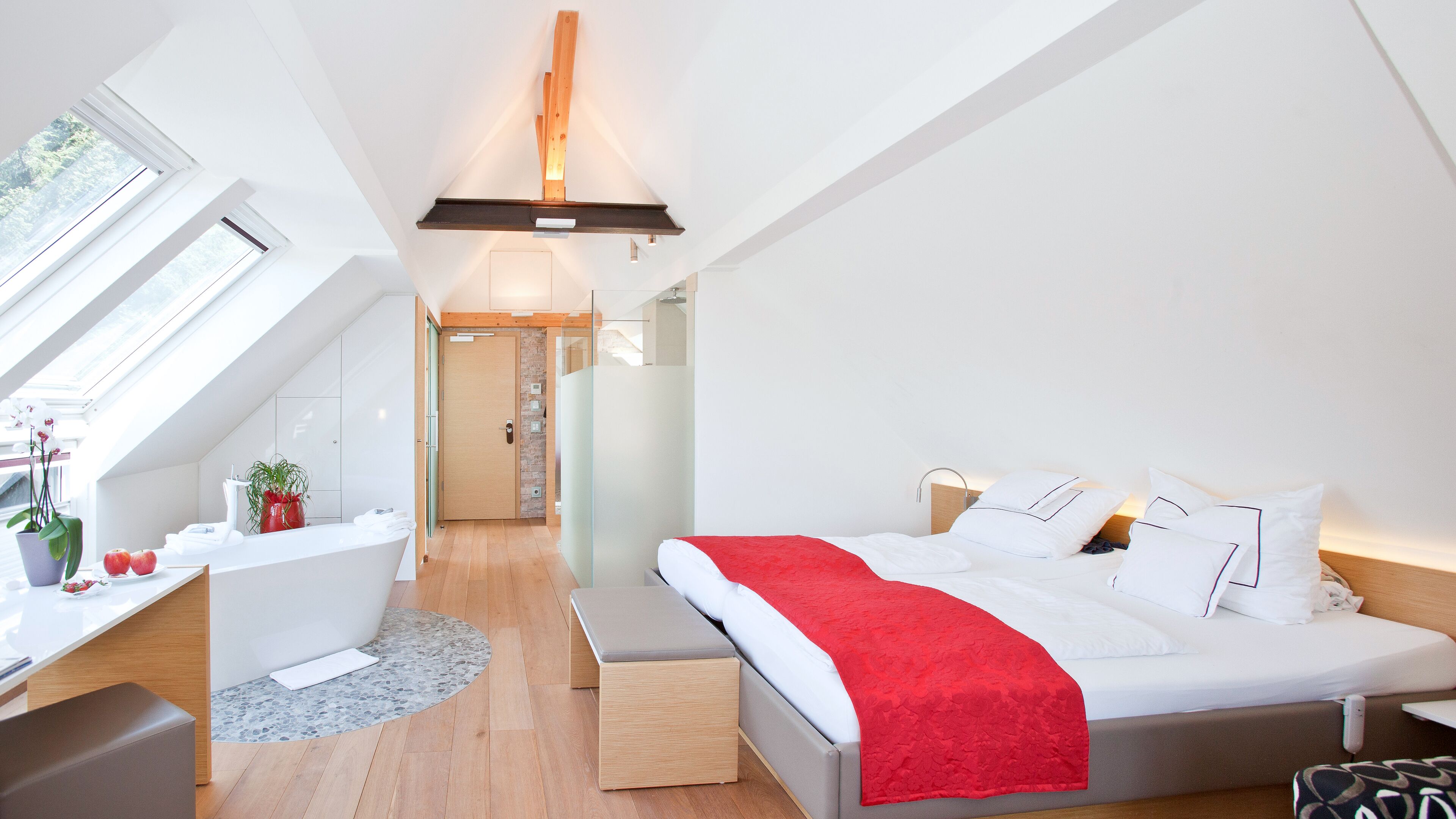Un dormitorio moderno con tragaluz, una cama con manta roja, una mesita de noche y un pequeño escritorio con una planta.