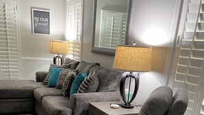 Un salon moderne avec un canapé d’angle gris, un fauteuil inclinable gris et une petite table avec une lampe. Un cadre décoratif est accroché au mur et un grand miroir se trouve au-dessus du canapé.