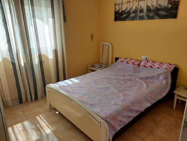 A bedroom with a bed, a nightstand, a chair, a window with curtains, and a painting on the wall.